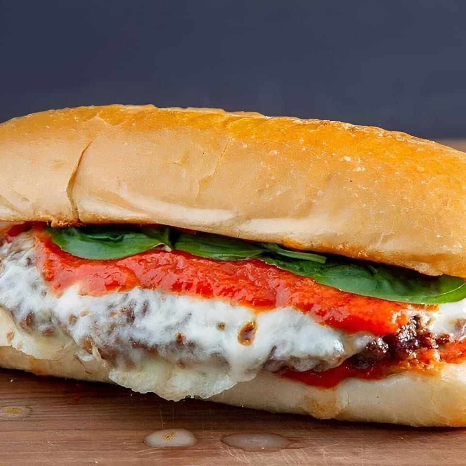 A close-up of a meatball sub with melted cheese, red sauce, and fresh basil on a wooden surface.