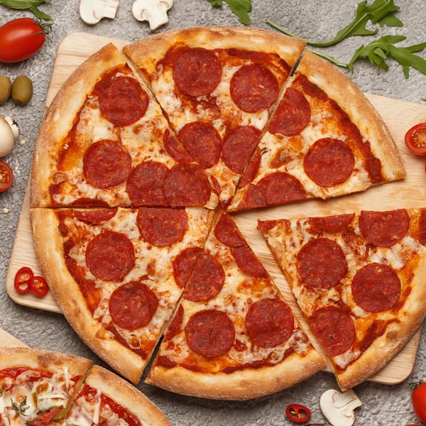Sliced pepperoni pizza on a wooden board, surrounded by fresh ingredients like tomatoes, mushrooms, and olives.