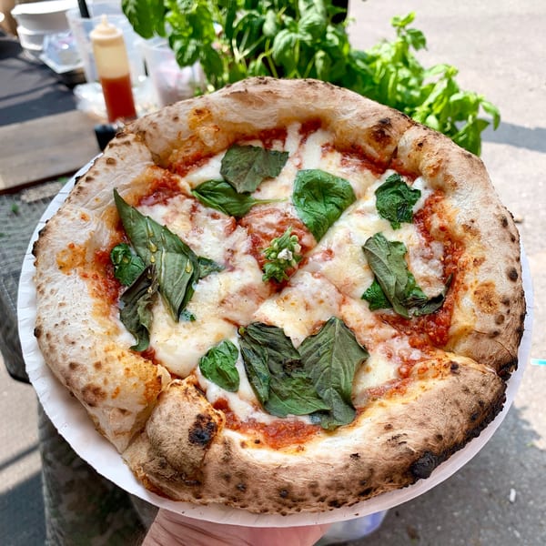 A hand holds a freshly baked Italian pizza with melted cheese, tomato sauce, and basil leaves.